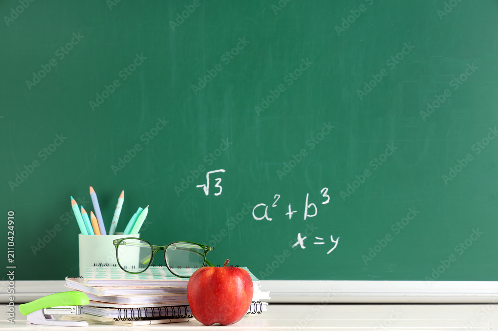 Obraz premium Apple, eyeglasses and stationery on desk near chalkboard with formulas. Back to school concept