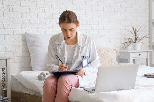 Female medical student studying with modern laptop and clipboard on bed at home