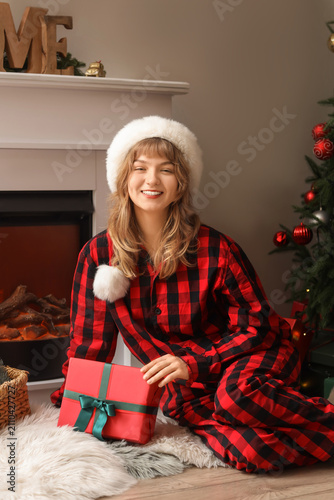 Pretty young woman in Santa hat with Christmas gift box sitting on floor at home