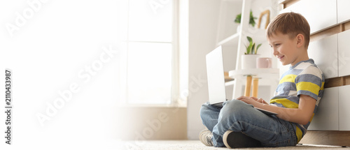 Smiling young boy using a laptop computer while sitting on the floor at home,...