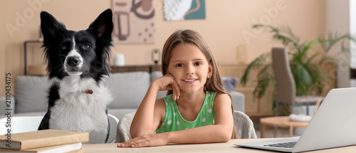 Smiling little girl and border collie dog sitting at a desk with a laptop and...