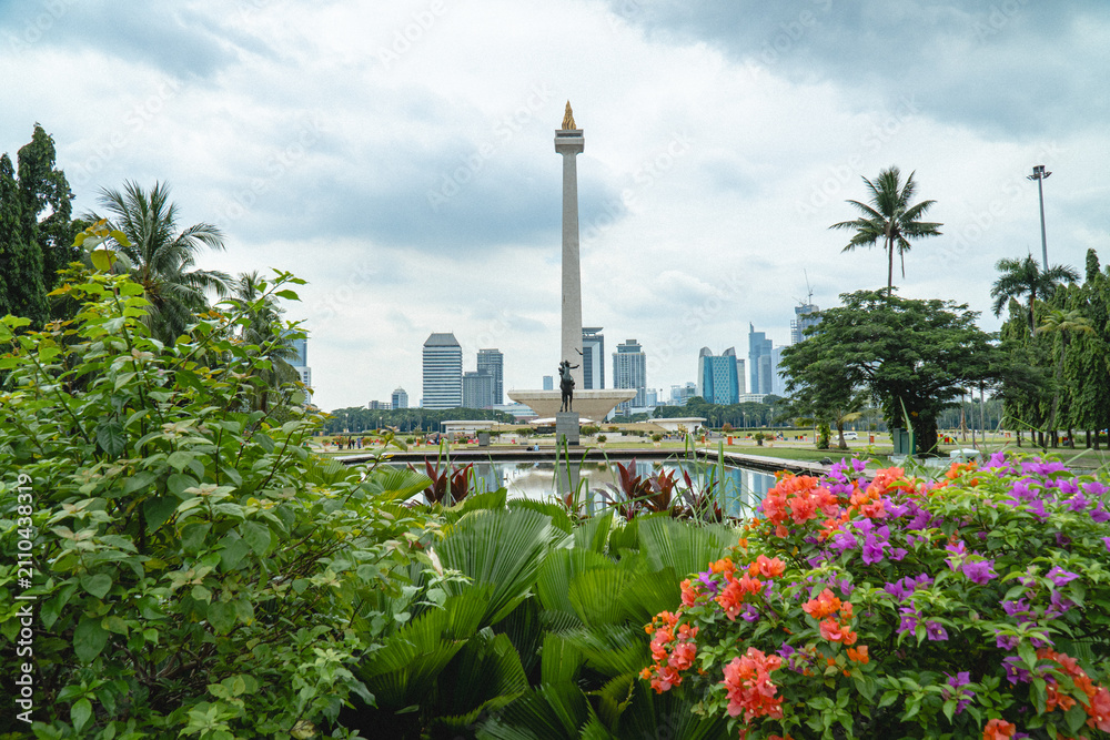 Obraz premium Jakarta, Indonesia - July 24, 2026: The National Monument (Monas) rises above landscaped gardens and colorful flowers at Merdeka Square, with Jakarta's skyline in the background.