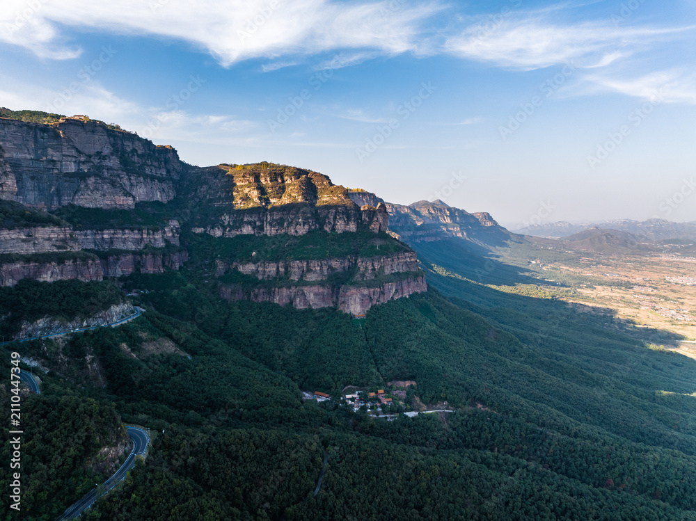 Fototapeta premium A panoramic landscape featuring layered rocky cliffs, dense green valleys, and a winding road under a clear blue sky. Small settlements nestle in the forested slopes.