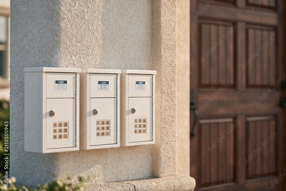 Fototapeta premium Modern Cream Mailboxes Attached to Textured Pillar Beside Rich Wooden Door with Sunlight