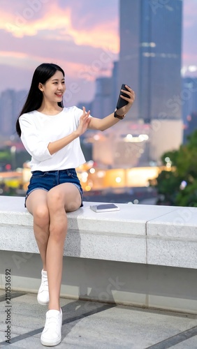 A young woman with dark hair takes a selfie outdoors. She wears a white top, shorts, and white sneakers, with a city skyline backdrop