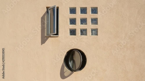Minimalist architectural facade with open window, glass block grid, and round porthole window casting soft shadows on beige wall.