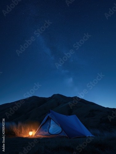 Small blue camping tent illuminated by a warm lantern under a starry night sky with visible Milky Way, set against rugged mountains and grassy terrain in serene wilderness.