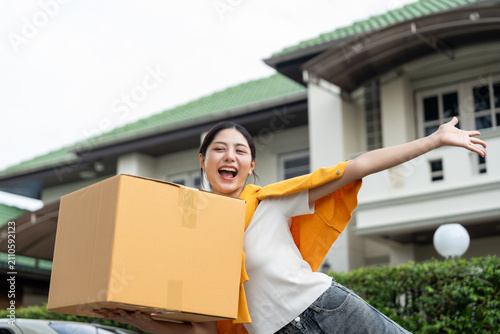Happy young woman holding moving box and house keys in front of new home. Moving day, real estate purchase, mortgage loan, home ownership and property investment concept.