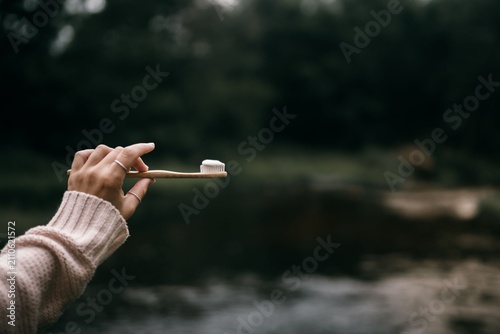 Fotografie Hand holds a wooden toothbrush with a dollop of white toothpaste against a blurred natural background of dark green forest foliage and a reflective river surface during a bright morning forest trip
