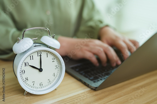 Working against clock, alarm clock on desk with person typing on laptop, concept of deadline, working hours, and daily routine.