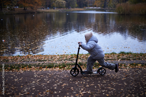 Child rides a scooter by a lake surrounded by trees during autumn with fallen leaves on the ground