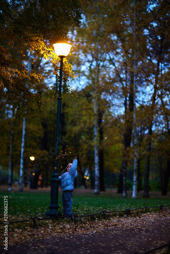 Boy reaches for a street light in a park during the evening among trees with yellow leaves