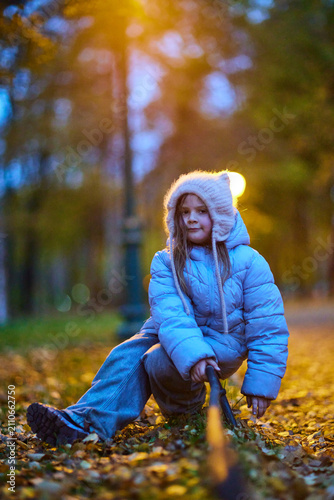 Child playing with a stick on a path in a park during evening hours surrounded by trees and street lights