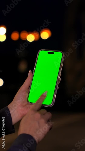Smartphone with Vertical Green Screen Chroma Key in Woman's Hands Against of Evening City Lights. Vertical Video