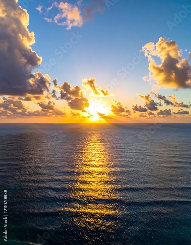 Aerial view of an ocean at sunset, showcasing golden sunlight reflecting onto the waves as clouds fill the sky. The image has a warm color scheme