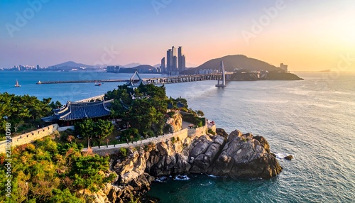 Aerial view of coastal city. A traditional building sits on a rocky outcrop. Skyscrapers, bridge, and calm sea blend with the dawn