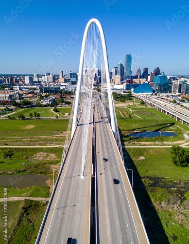 Aerial view of a sleek, white arch bridge spanning a highway, with a city skyline and park in the background
