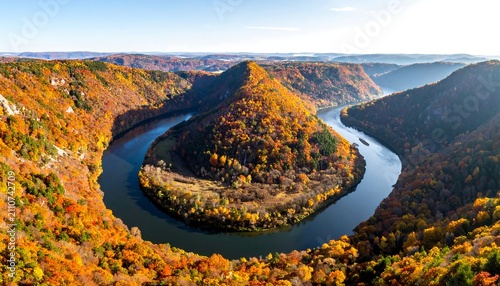 Aerial view captures a striking river bend, encircled by vibrant autumn foliage, under a bright blue sky. Sunlight bathes the landscape