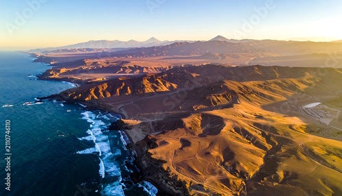 Aerial view of coastal landscape with ocean waves crashing against a rugged shoreline, hills and mountains in the distance, bathed in golden sunlight