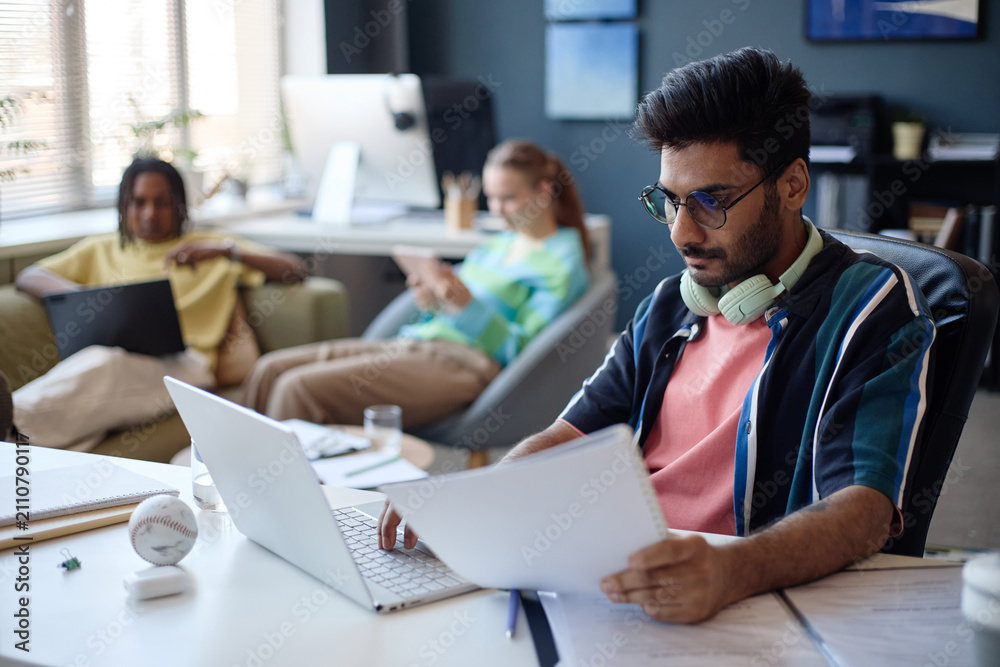 Fototapeta premium Young man working on laptop and reviewing documents in shared office while colleagues using devices in background, representing teamwork and productivity. For startup marketing or workplace content
