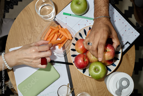 Obraz na plátne Overhead shot of adult coworkers sharing apples and carrot sticks over work documents, promoting healthy office snacking and wellness