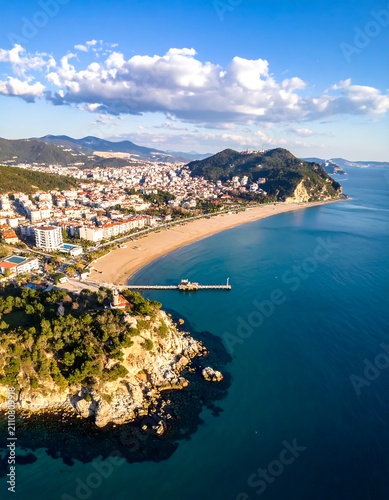 Aerial view of a coastal city, showcasing a long sandy beach, turquoise water, and a scenic mountainous background