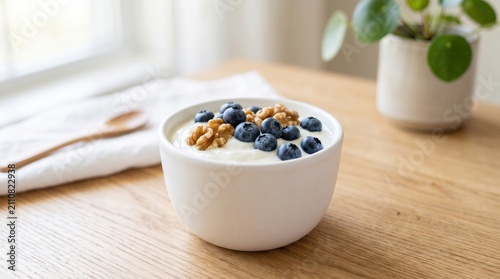 A white bowl of yogurt topped with blueberries and walnuts sits on a wooden table next to a wooden spoon and a potted plant by a window.