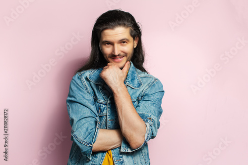 Young happy man with long hair holding hand under chin. Isolated on pastel pink background.