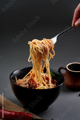 Hand twisting spaghetti bolognese with a fork over a black bowl on dark background. Action food shot.