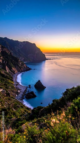 Aerial view of a coastline at dawn. Golden sunlight illuminates a calm sea and beach below rugged cliffs covered in greenery