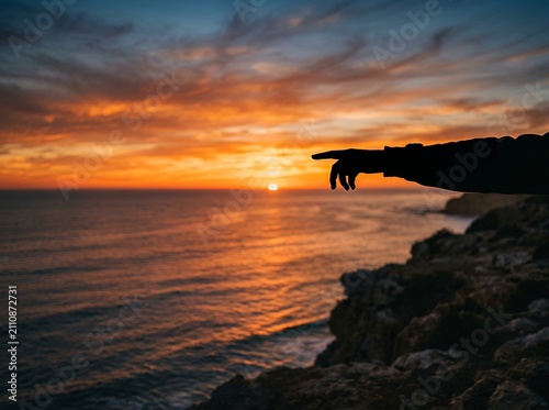 Silhouette of a hand pointing towards a dramatic ocean sunset over rocky cliffs.