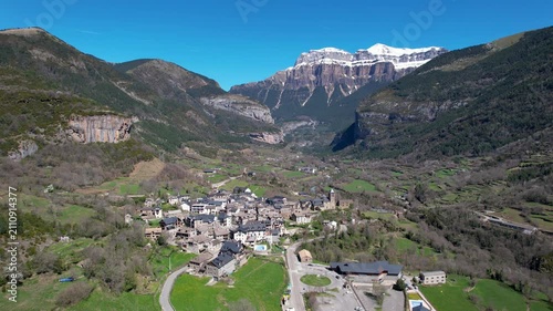 Panoramic aerial shot of Torla-Ordesa village and covered with snow mountains of Ordesa y Monte Perdido National Park on sunny spring day. Huesca, Aragon, Spain.