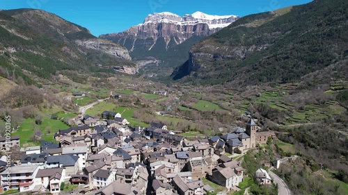 Backward reveal aerial shot of Torla-Ordesa village and covered with snow mountains of Ordesa y Monte Perdido National Park on sunny day. Huesca, Aragon, Spain.
