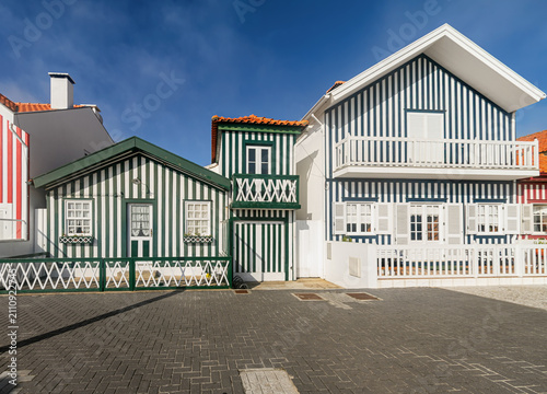 Traditional striped houses with wooden facades, balconies and shutters stand beneath a clear blue sky in Costa Nova, Portugal.