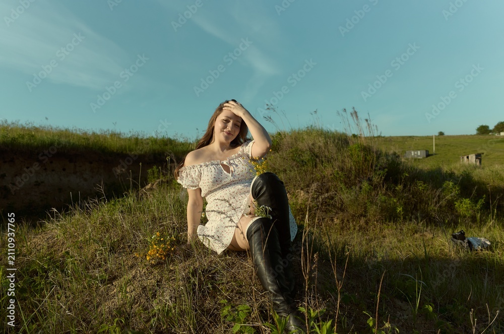 Obraz premium Model sits on a hill covered in grass and flowers while wearing a white dress and black boots during sunny weather