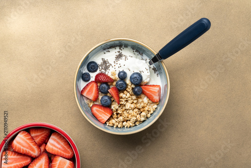 Creamy yogurt topped with crunchy granola, strawberries, blueberries and chia seeds served in a ceramic bowl on a warm beige background with copy space.