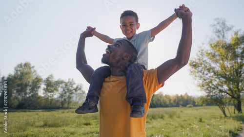 Father carries his son on his shoulders while they laugh and play in a green field at sunset. Quality time spent together in nature.