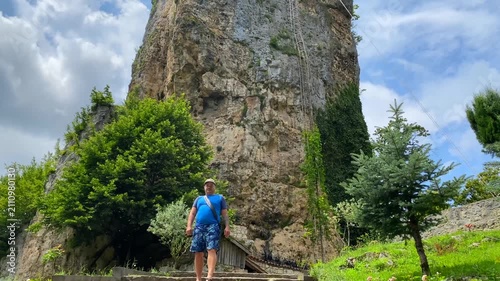 A traveler explores the landscaped grounds beneath Katskhi Pillar, where a secluded monastery crowns a towering limestone monolith, highlighting faith, heritage, geology, and rural tourism in Georgia