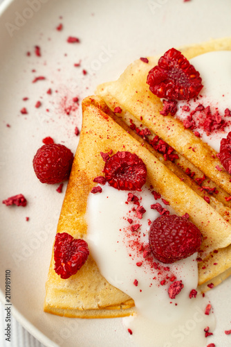 Crepes are arranged on a white plate. Fresh raspberries and cream are placed on top. Red raspberry powder is sprinkled around the crepes. It is a bright morning setting