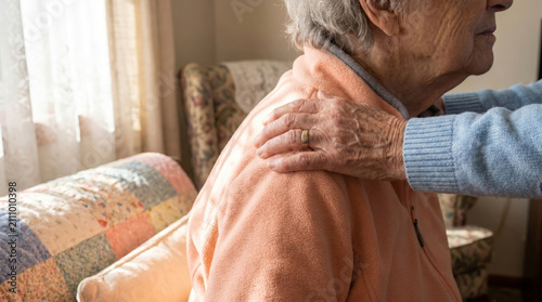 Caregiver hand resting on shoulder of elderly woman in nursing home. Compassionate support and palliative care for senior. Comforting touch in hospice setting for aging patient.