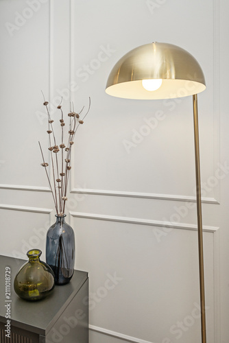 Elegant brass dome floor lamp beside glass vases with dried branches, styled against paneled white wall in minimalist interior.