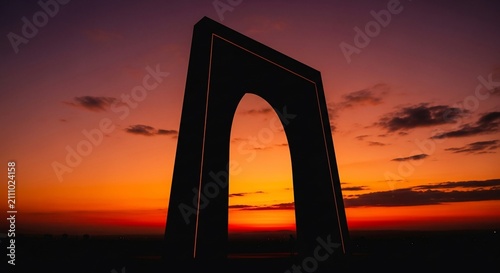 Architectural arch stands silhouetted against a brilliant orange sunset sky during the late evening hours.