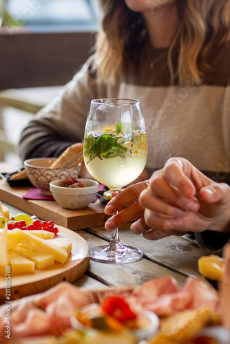 Blonde female hand with white cocktail glass during aperitivo on table with cold cuts and cheeses