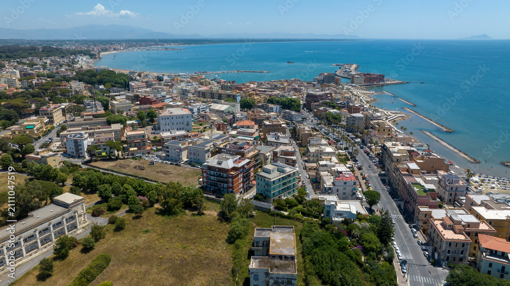 Fototapeta premium Aerial panoramic view of the sunny promenade and coastline in Anzio, in province of Rome, Italy. There are beachfront buildings, sandy shores, breakwaters, and clear sea under a bright summer sky.