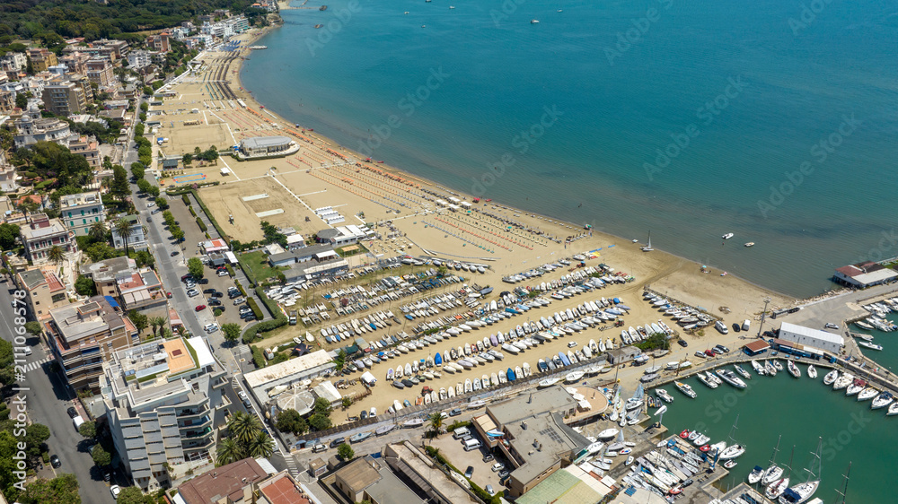 Fototapeta premium Aerial view of the coast of Anzio, in Lazio, Italy, featuring a wide sandy beach, a boat dry dock storage area, a circular harbor filled with yachts, and beachfront buildings along the clear blue sea.
