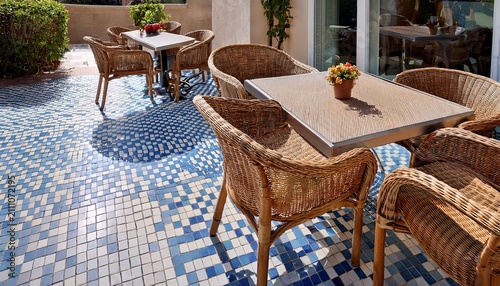 upscale outdoor restaurant patio features woven rattan chairs and light oak dining tables placed nicely upon blue and white checkered tiles