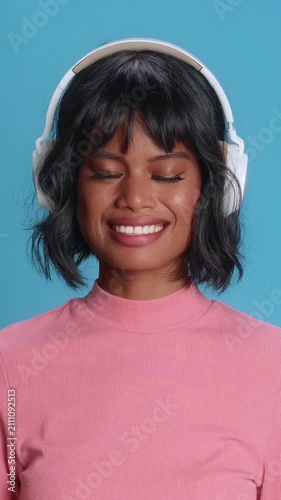 Attractive Asian woman with short hair listens to music with white headphones, smiling with her eyes closed, studio footage captures a peaceful and happy mood on blue background 