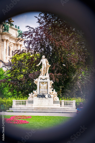 Vienna, Austria - June 9, 2023: Mozart monument in Burggarten Vienna with white marble statue, ornate pedestal, lush green foliage, historic building facade, and colorful flower beds