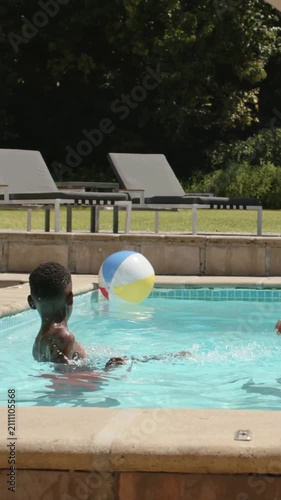 Vertical video: Chasing youth in red trunks kicking drifting beach ball in home pool, adult joining