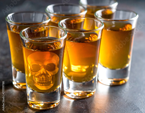 Multiple shot glasses filled with strong amber liquor with a subtle skull reflection.